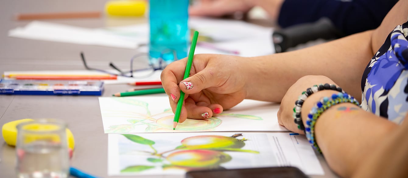 Closeup of a woman's hands as she uses watercolor pencils to sketch fruit on a branch.