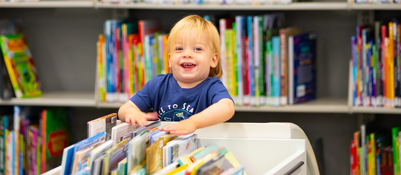 A toddler offers a wide smile to the camera while rifling through a book bin.