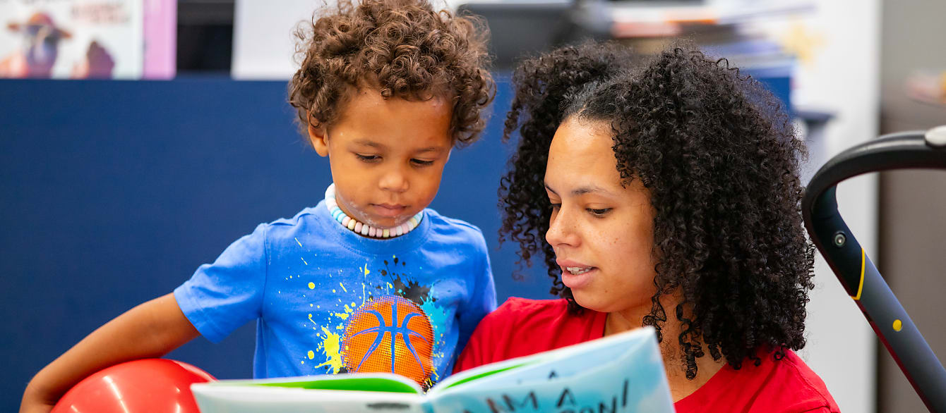 A toddler holding a red balloon looks intently at a book while his mother reads to him.