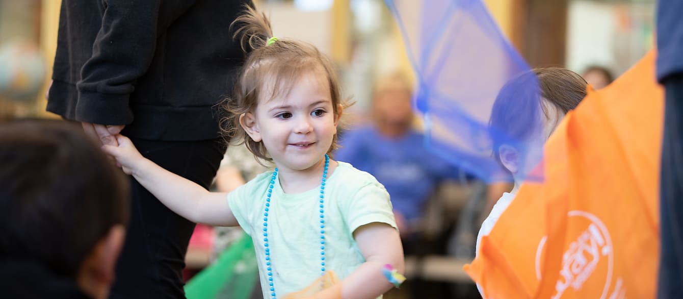 A preschool-age girl wearing a blue beaded necklace smiles as children holding tulle dance around her.