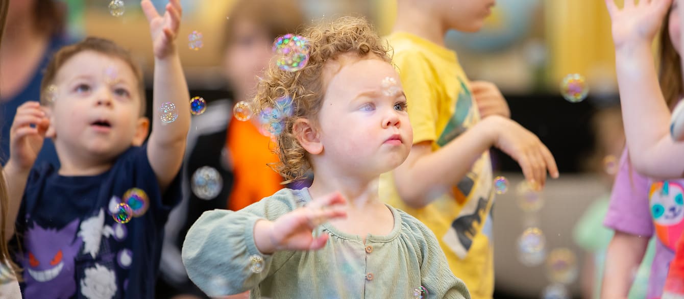 A toddler with curly blonde hair wearing a green shirt stares at the bubbles floating around her.