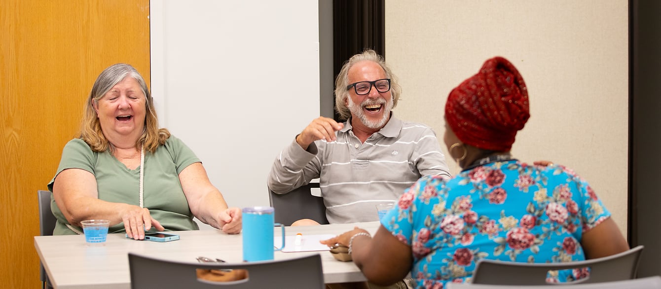 A group of three adults sits around a table, laughing while holding small white boards.