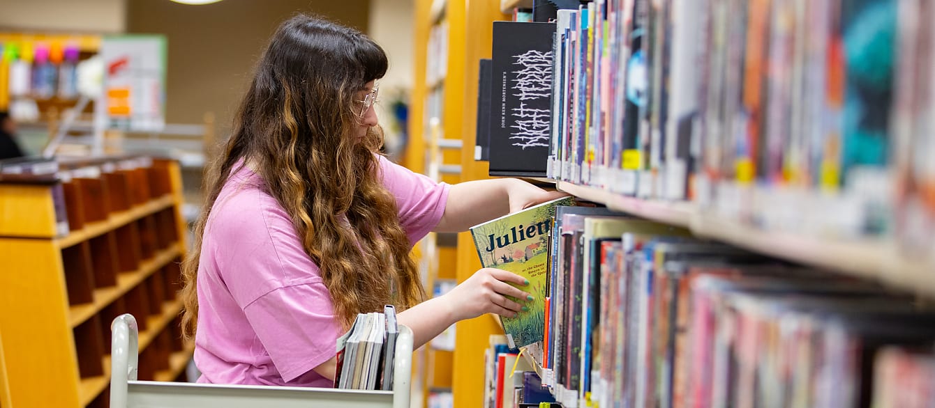A librarian in a bright pink shirt shelves a book, while a cart sits in the foreground.