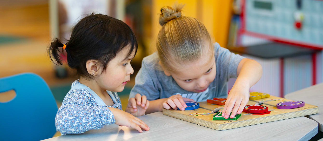 A little girl watches as a little boy works on a wooden puzzle.