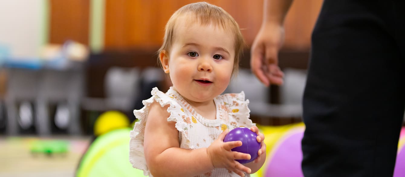 A baby in a frilly tank top, holding a purple plastic ball smiles at the camera.