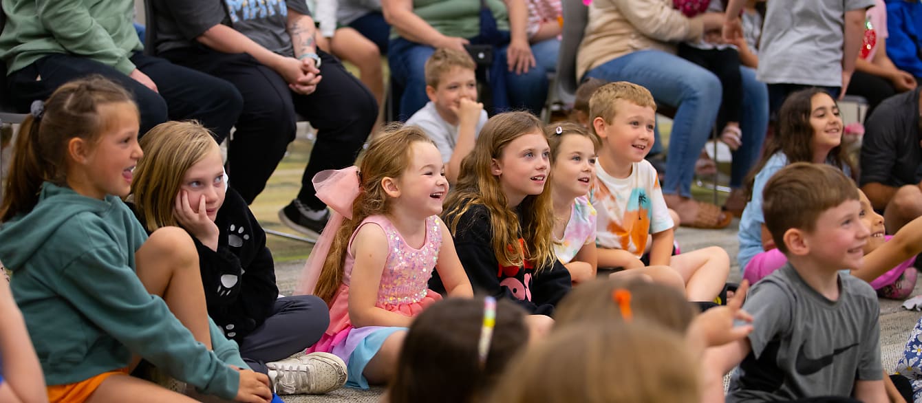 A crowd of children sit on the floor smiling as they watch a magic show.