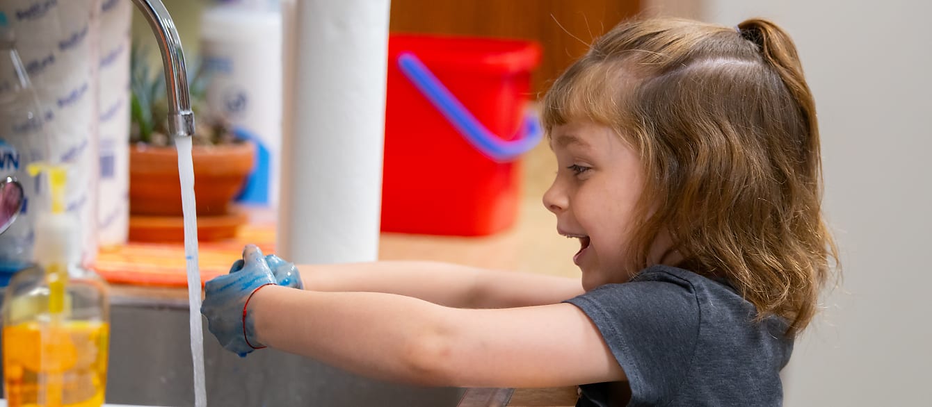 A school-age girl rinses her hands, covered in blue paint, under a faucet.