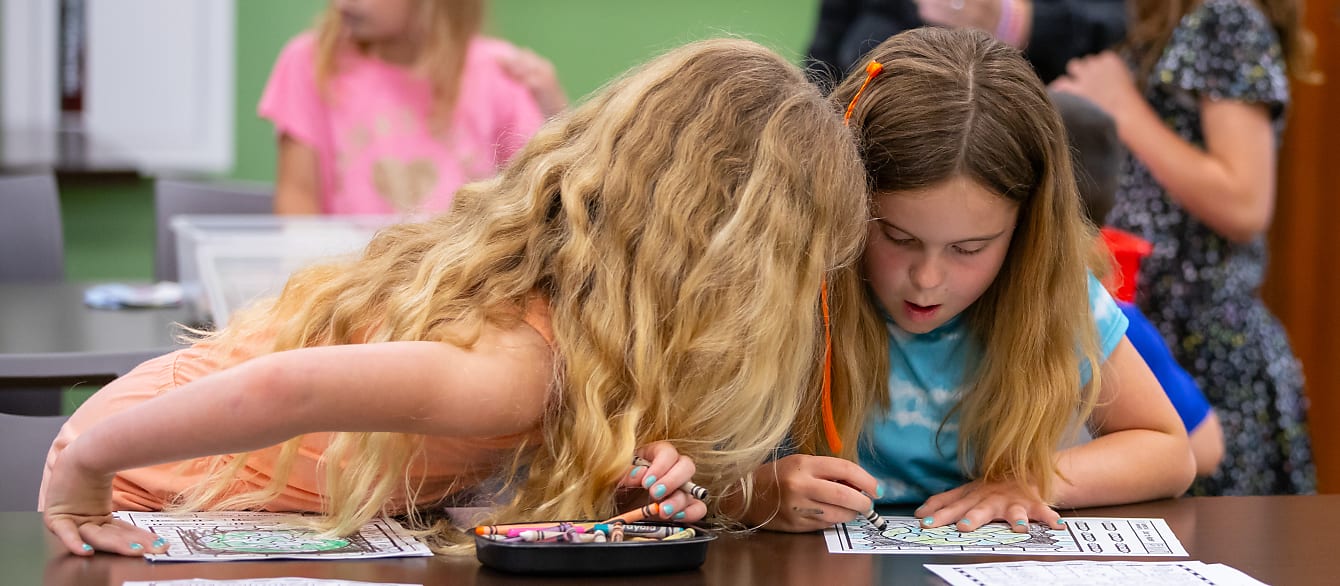 Two middle-school girls color together. The one on the left is leaned over watching the one on the right color.