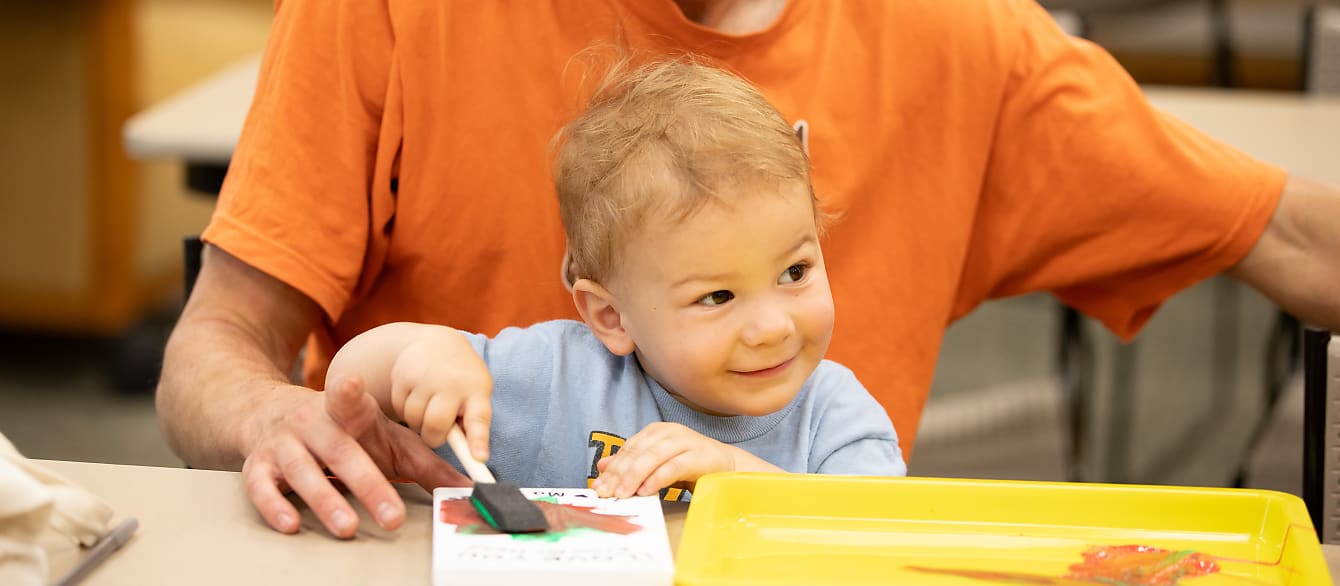 A toddler sitting on his father's lap uses a sponge brush to paint with red and green on a canvas.