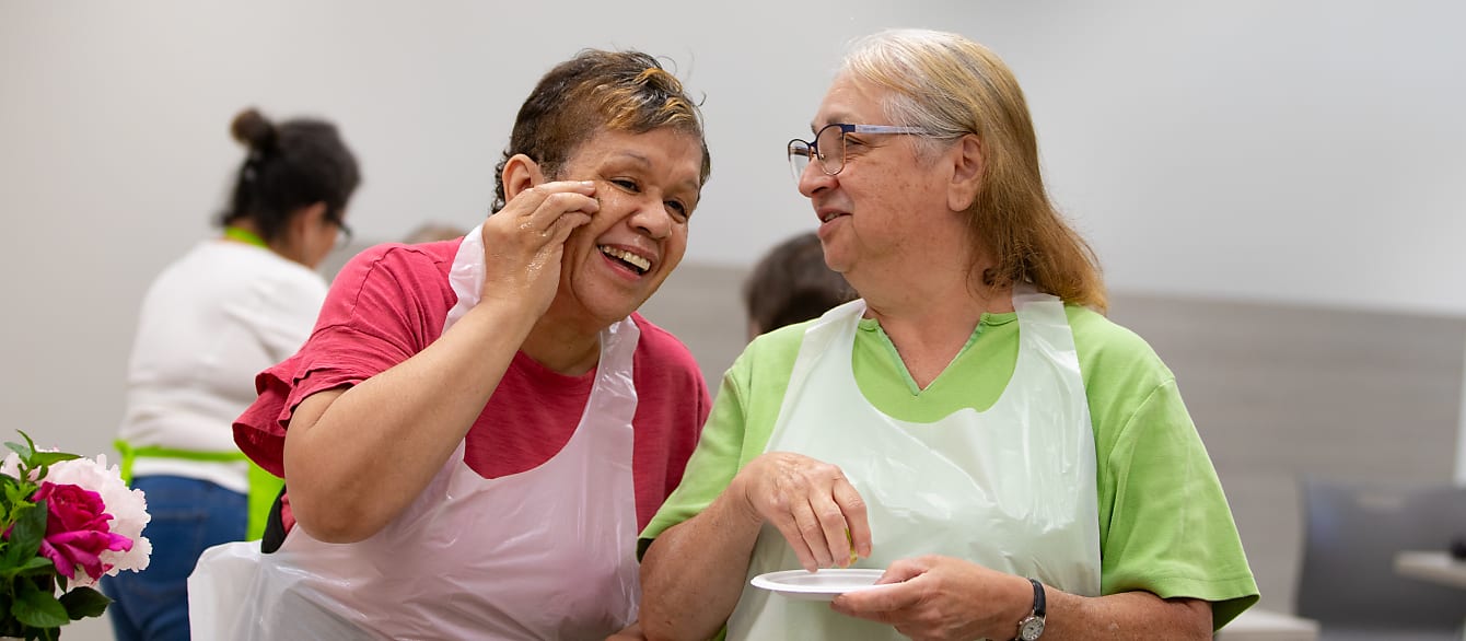 A woman in a pink shirt laughs with a woman in a green shirt as they experiment with natural skin care.