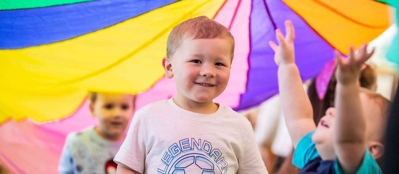 A preschool-age boy smiles at the camera while standing underneath a multicolored parachute.