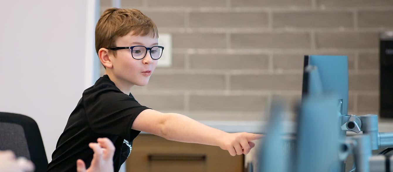 A young boy wearing black-frame glasses points excitedly at a computer screen.
