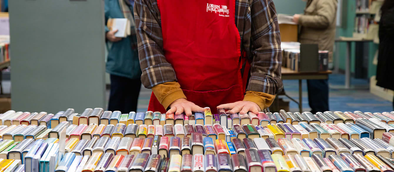 In the foreground, rows and rows of book spines. In focus at the end of the frame, a man wearing a bright red apron organizes