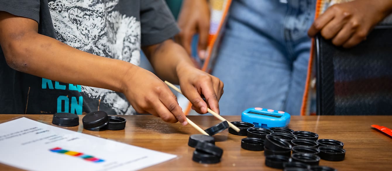 A close up of a boy trying to pick up bottle caps and stack them using chopsticks