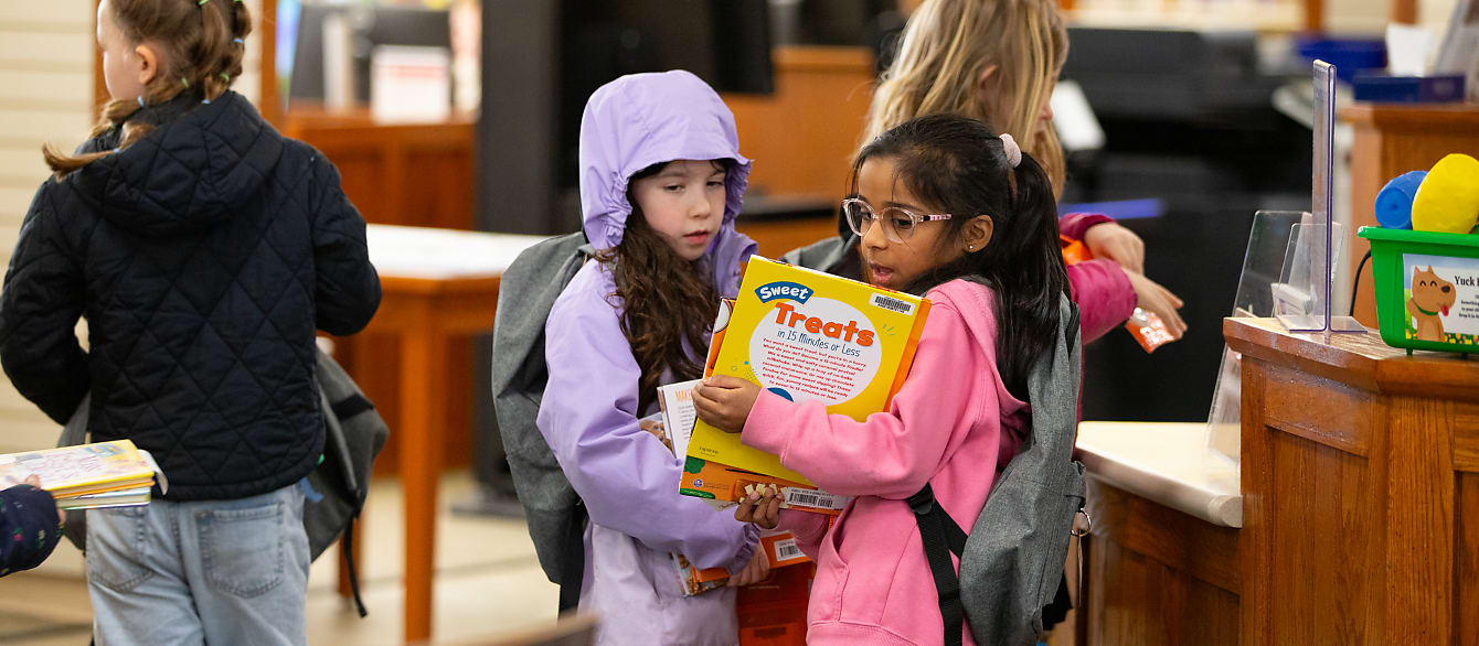 Two young girls in light coats look at a book while standing in line to check out.