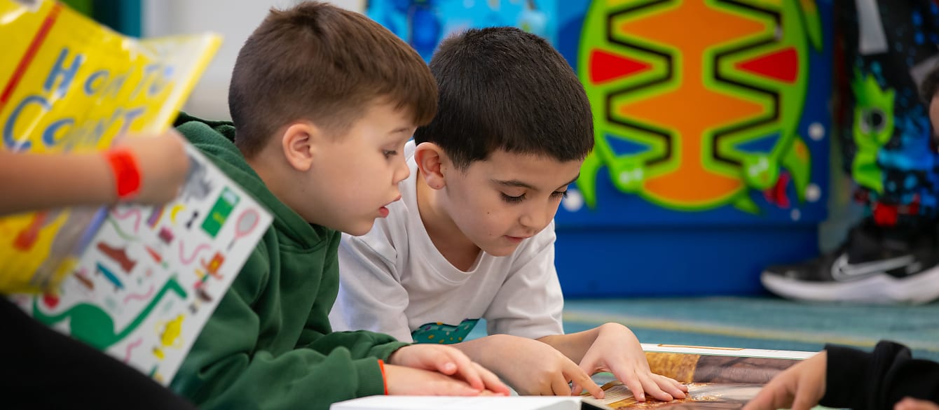 Two little boys lay on their stomach reading together in front of a colorful background.