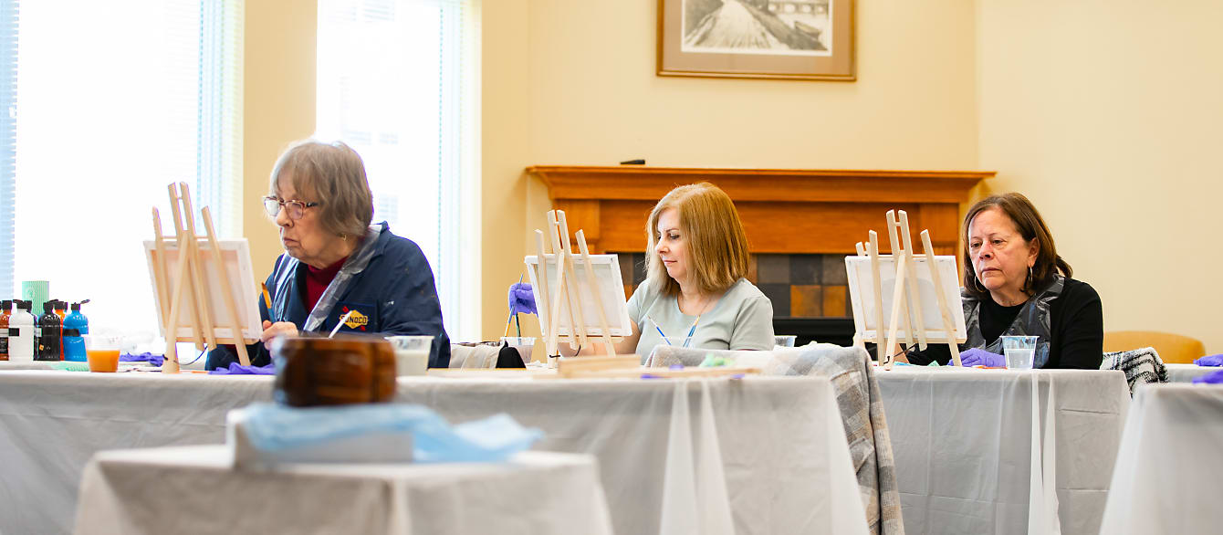 Three women each paint on a small canvas directly in front of them