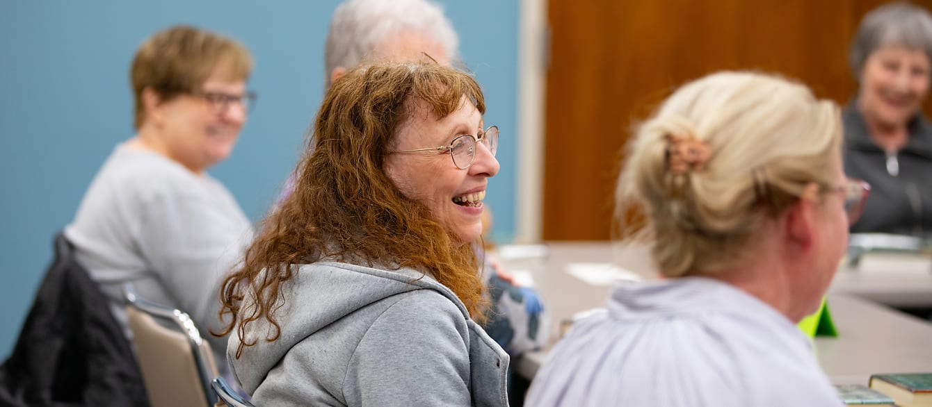 A group of women sit around a table and laugh. One is in focus in the middle, the rest are out of focus.