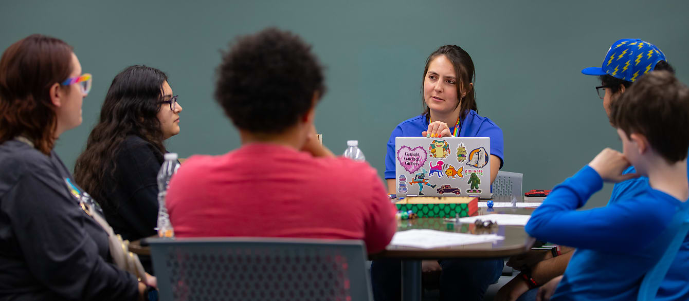 A group of teenagers sit around a table while a librarian narrates their game environment.