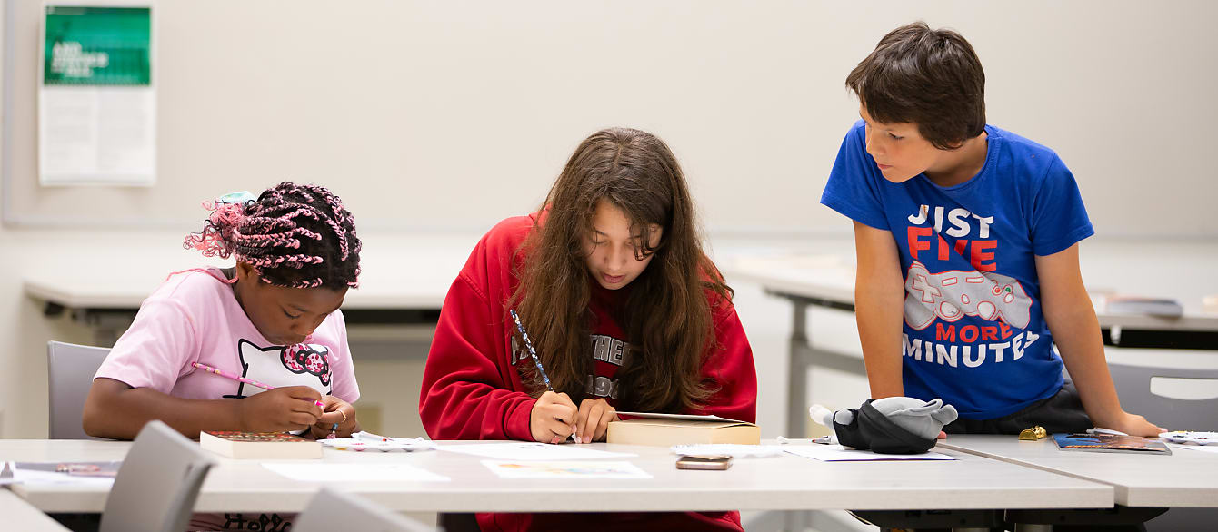 Two young tweens on the left side of the table bedazzle books while a tween boy stands on the right and watches.