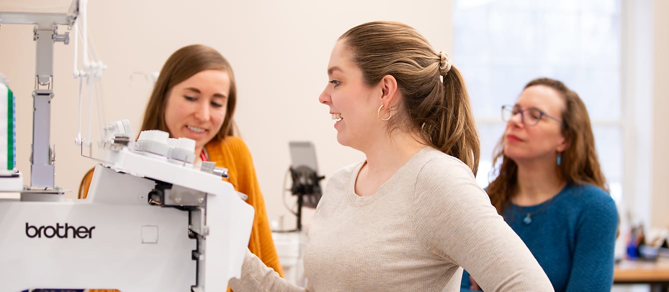 A woman in the foreground uses the touch screen on a large embroidery machine while two librarians help in the background