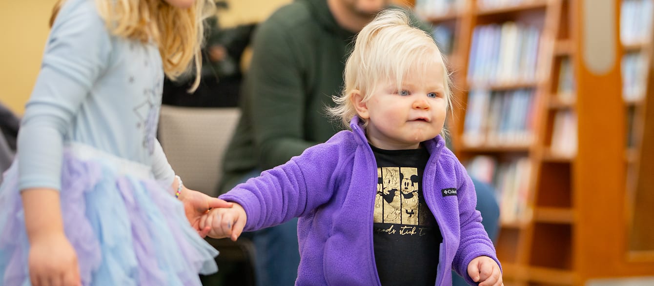 A young sister holds on to her baby sisters hand and helps her walk.