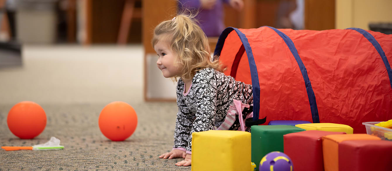 A toddler pokes her head out of a colorful tube surrounded by multicolored blocks and balls