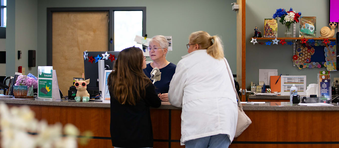 A woman in a white coat and a girl in a black coat check out a book from a librarian