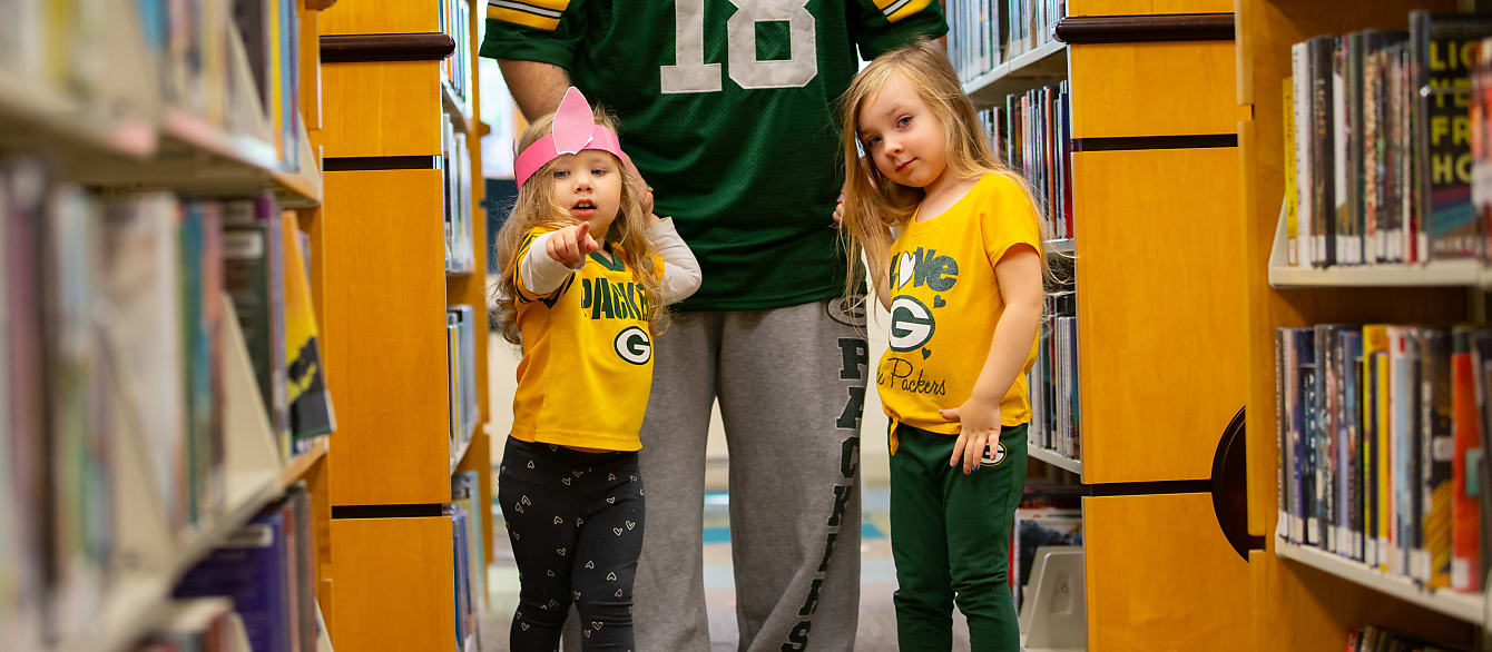 Two sisters wearing Green Bay Packers gear walk through books while holding dad's hand