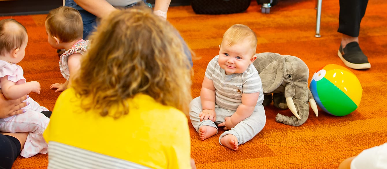 A baby sits on an orange carpet and smiles at his mother, blurred in the foreground.
