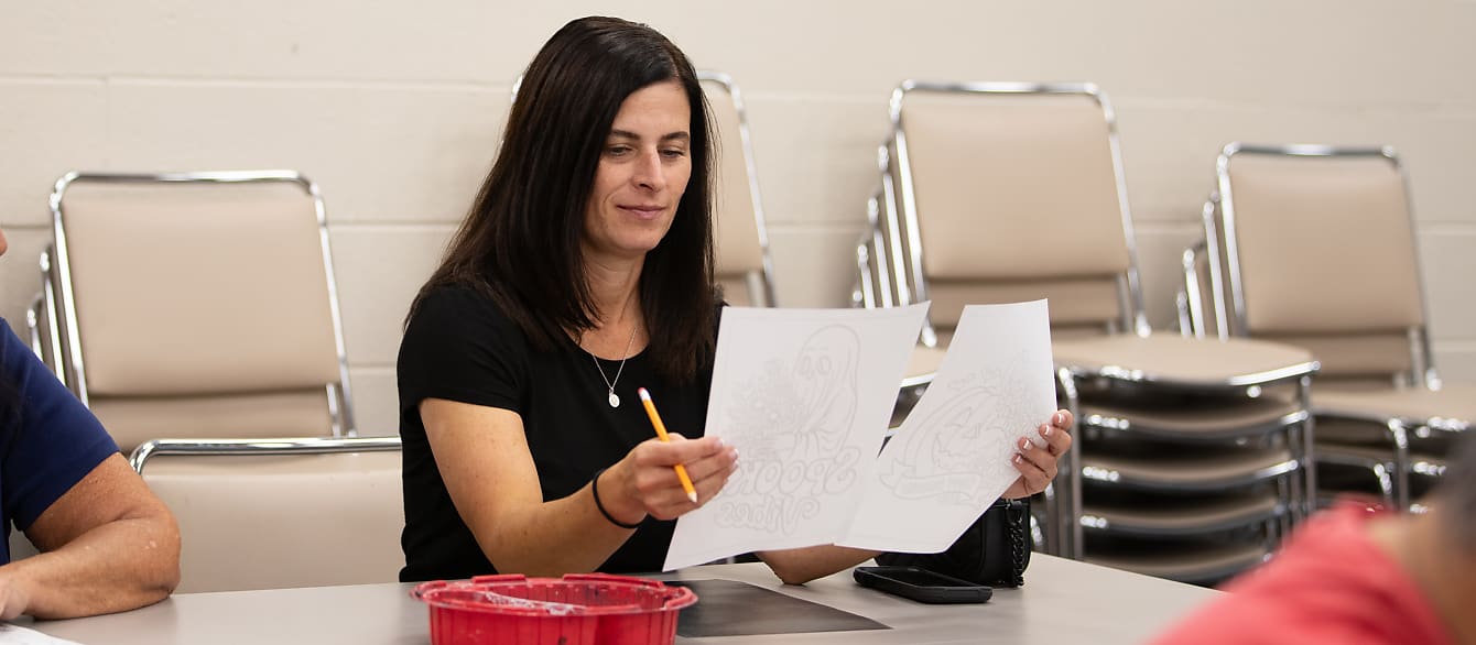 A woman compares two coloring pages; one of a cartoon ghost and one of a cartoon jack-o-lantern