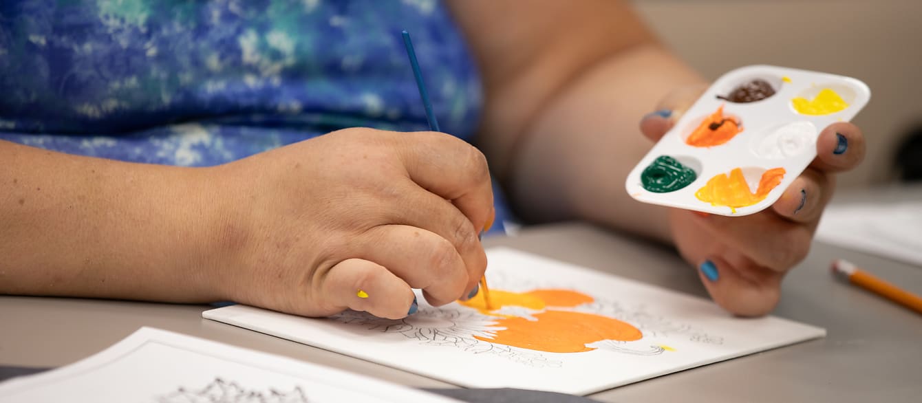 A closeup of a woman in a blue shirt painting a pumpkin on a canvas