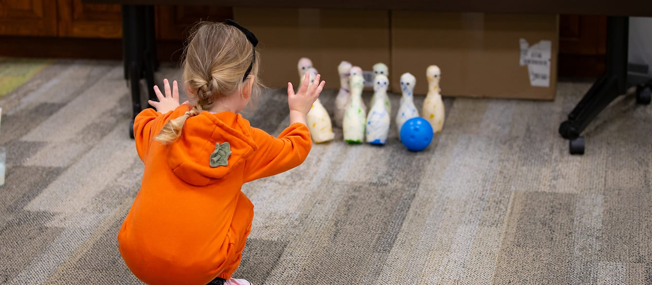 A young girl in a pumpkin onesie squats on the ground and throws a ball towards bowling pins decorated as mummies