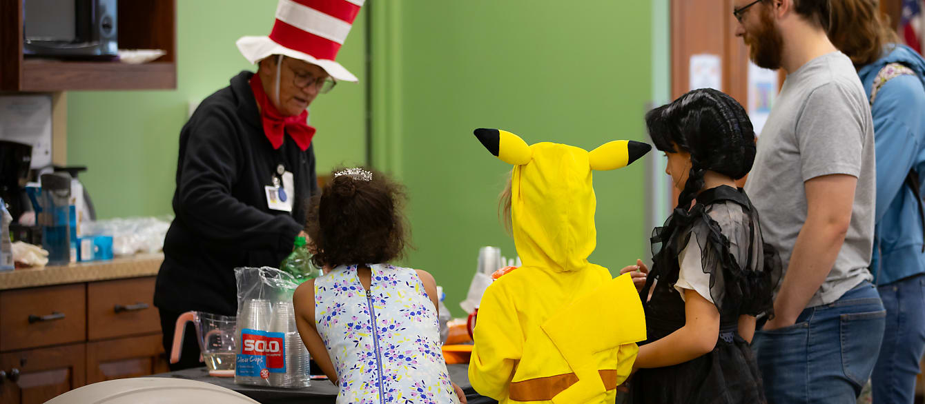 A librarian in a Cat in the Hat costume pours drinks for a girl in a Pikachu costume and a girl in a Wednesday costume.