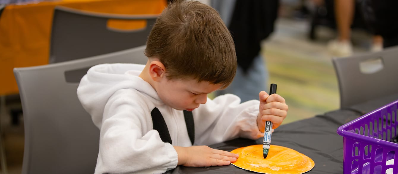 A young boy in a ghost onesie colors a paper pumpkin.