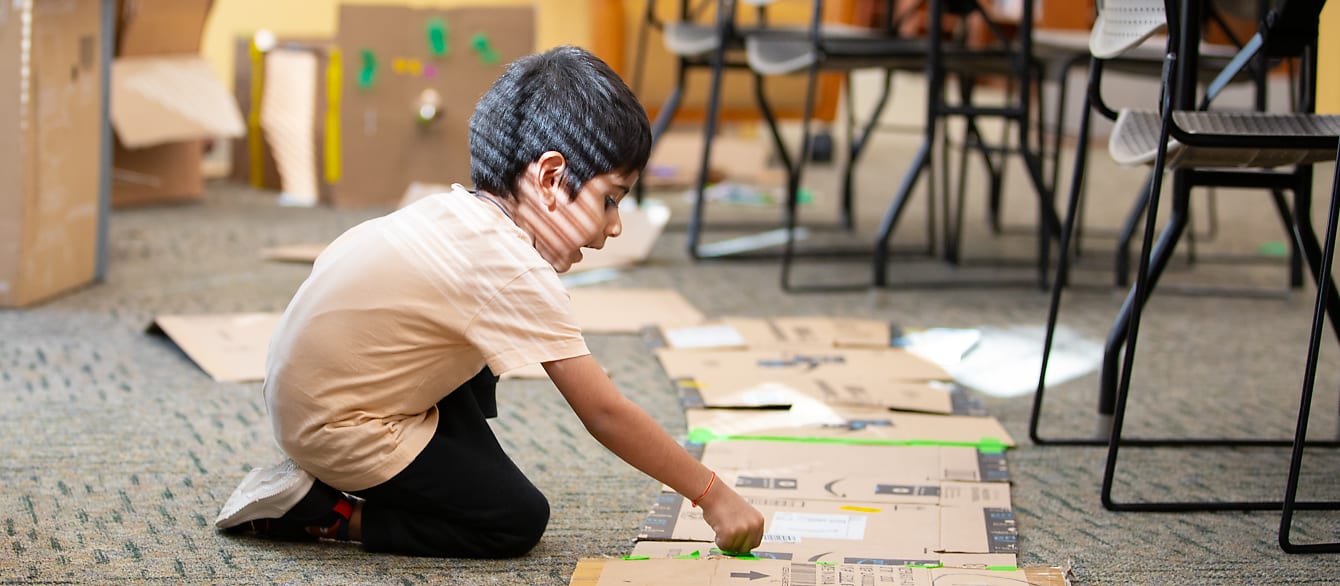 A young boy leans over a strip of cardboard with tape while sun from the window streams over his back.