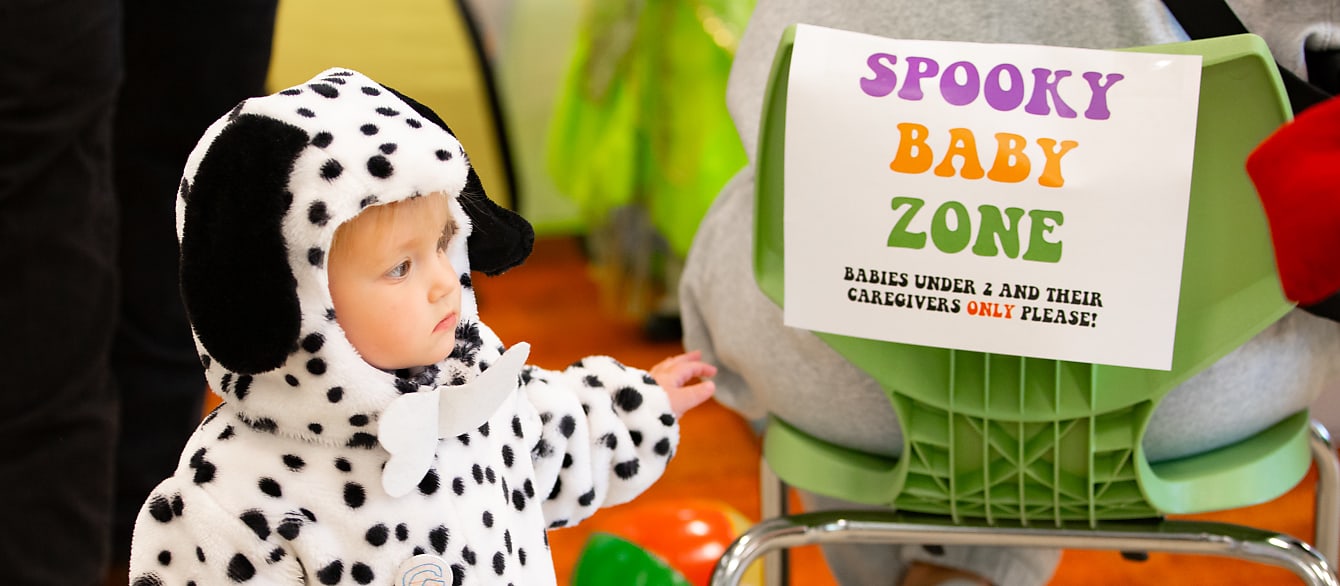 A toddler in a dalmatian costume stands beside a colorful sign that reads: "Spooky Baby Zone"