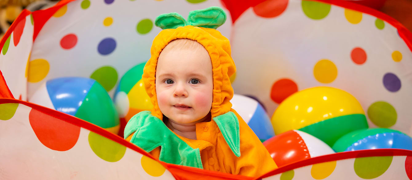 A baby in a pumpkin costume sits in a ball pit and smiles at the camera