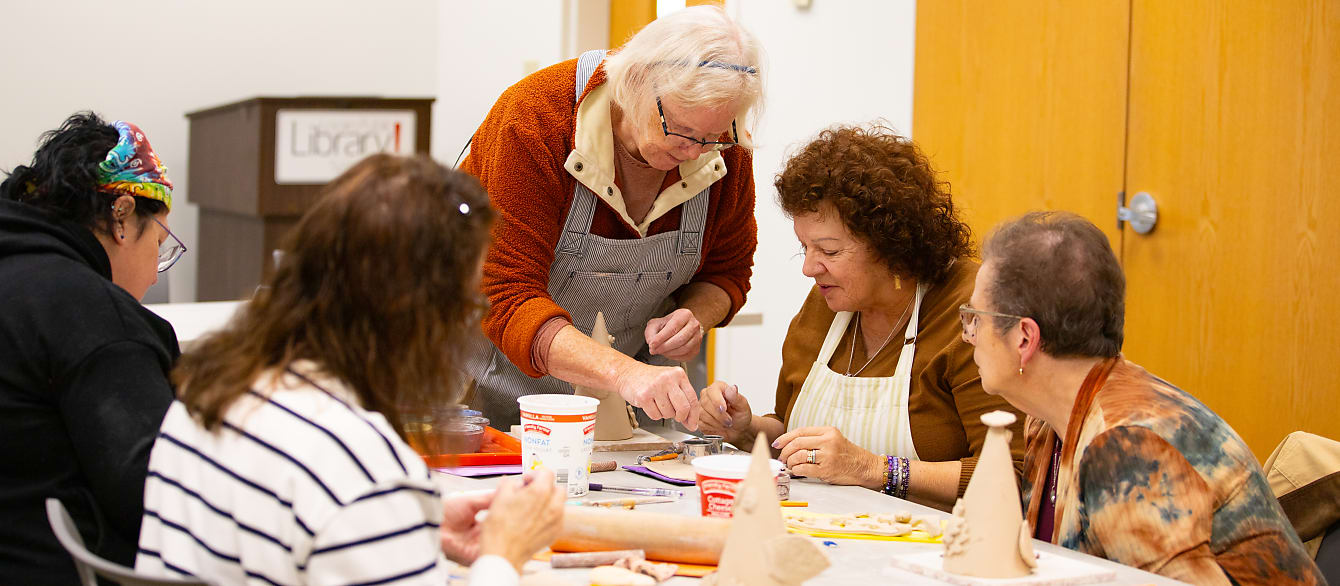 A group of women sit around a table covered in clay and sculpting material. The instructor is showing them how to sculpt.