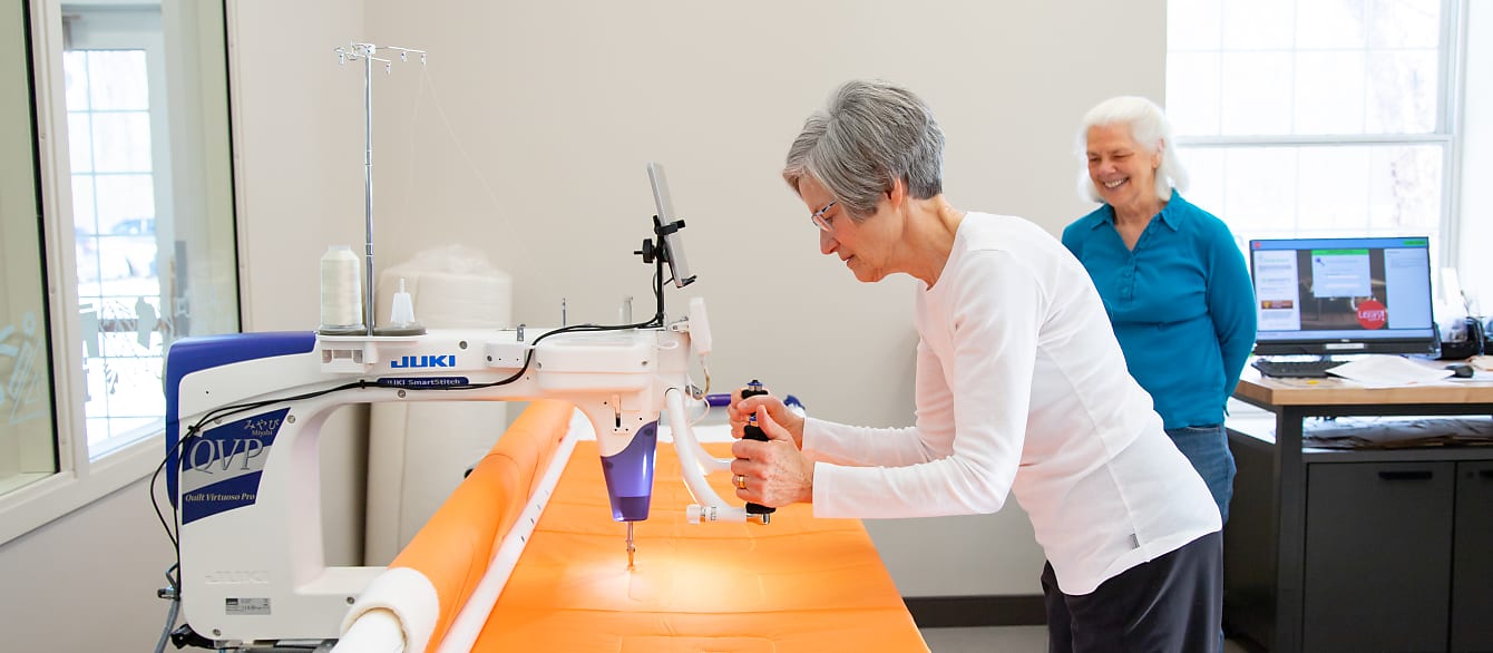 One woman operates a long arm quilting machine on bright orange fabric as another woman watches in the background.