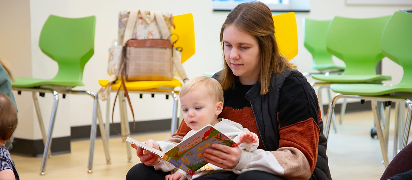 A mother sits on the floor with a child in her lap. Both of them are looking at the board book in her hands.
