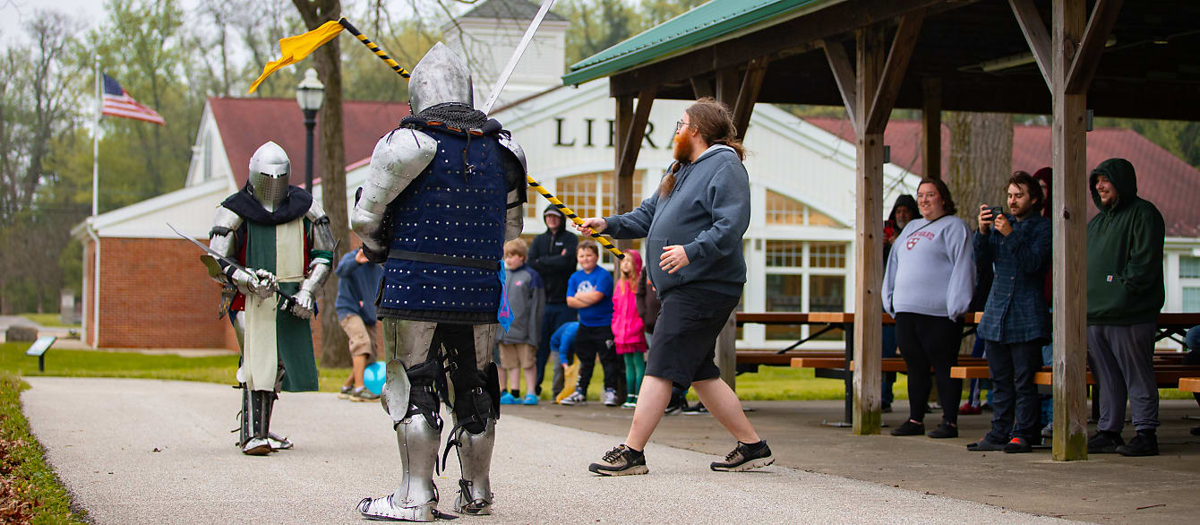 A crowd stands around two people dressed in knight armor fighting with swords