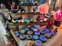 Colorful, glowing resin domes displayed on wood slices at a craft fair table, with other handmade goods and people in the background.