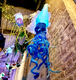 A large, blue squid sculpture with glowing lights hangs alongside other sea creature decorations in an indoor space with exposed brick walls and string lights.