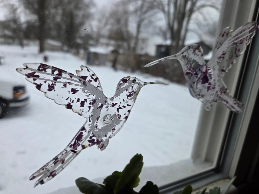 Two decorative translucent hummingbird stickers are attached to a window. Snow-covered ground and trees are visible outside.