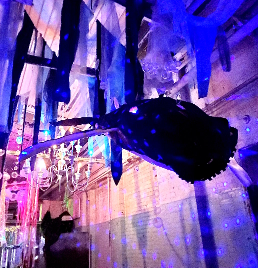 A dark whale sculpture hangs from a ceiling decorated with fabric strips and illuminated by purple and blue lights in an indoor space.